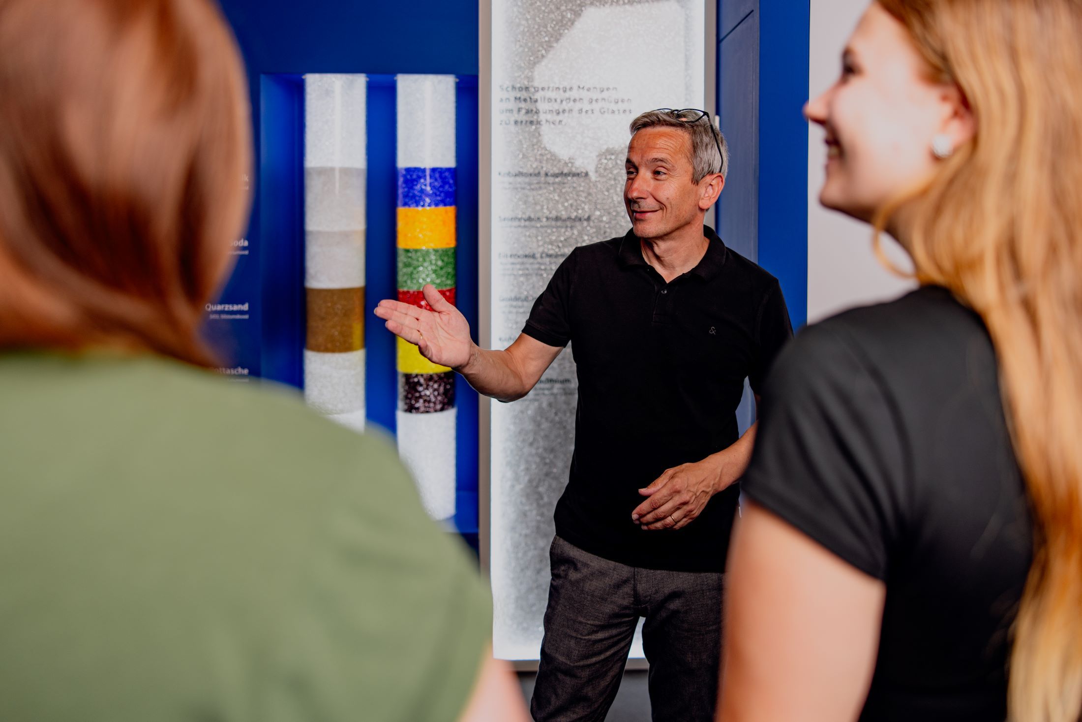 Ein Mann erklärt zwei Frauen etwas in einem Ausstellungsraum mit bunten Glasröhren.