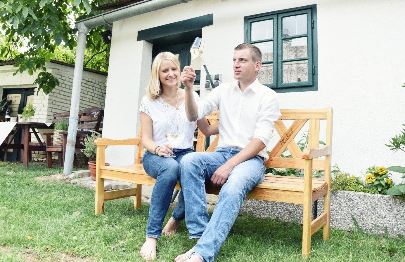 Ein Paar sitzt auf einer Holzbank vor einem Haus und stößt mit Weingläsern an.