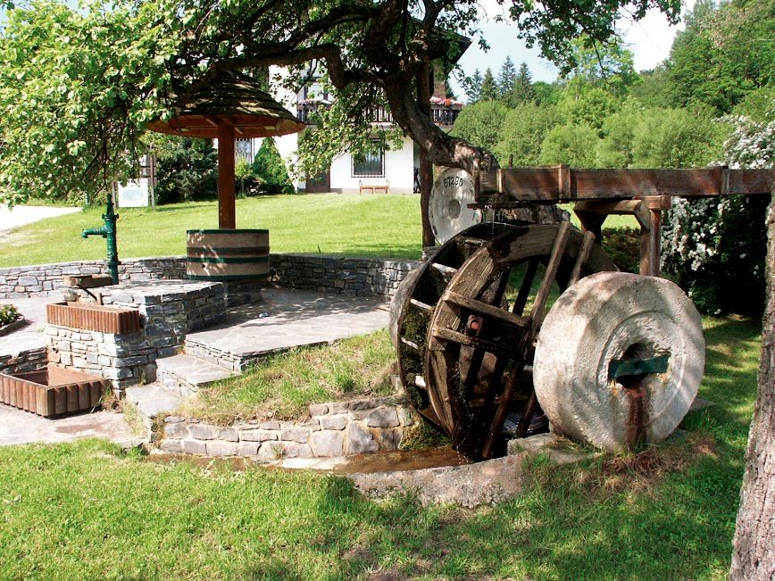 Altes Mühlrad mit Mühlstein, daneben Stufen und ein Brunnen, umgeben von Wiese und Bäumen.