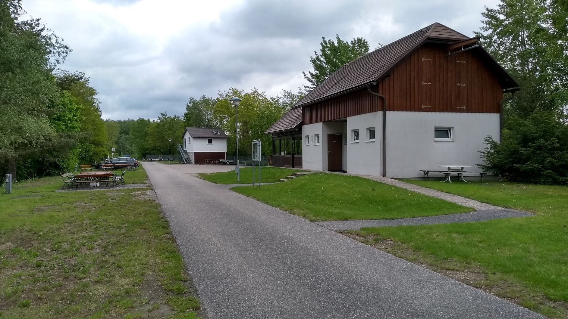 Ein Weg führt an einem weißen Gebäude mit Holzverkleidung vorbei, umgeben von Bäumen und Wiesen, unter einem bewölkten Himmel.