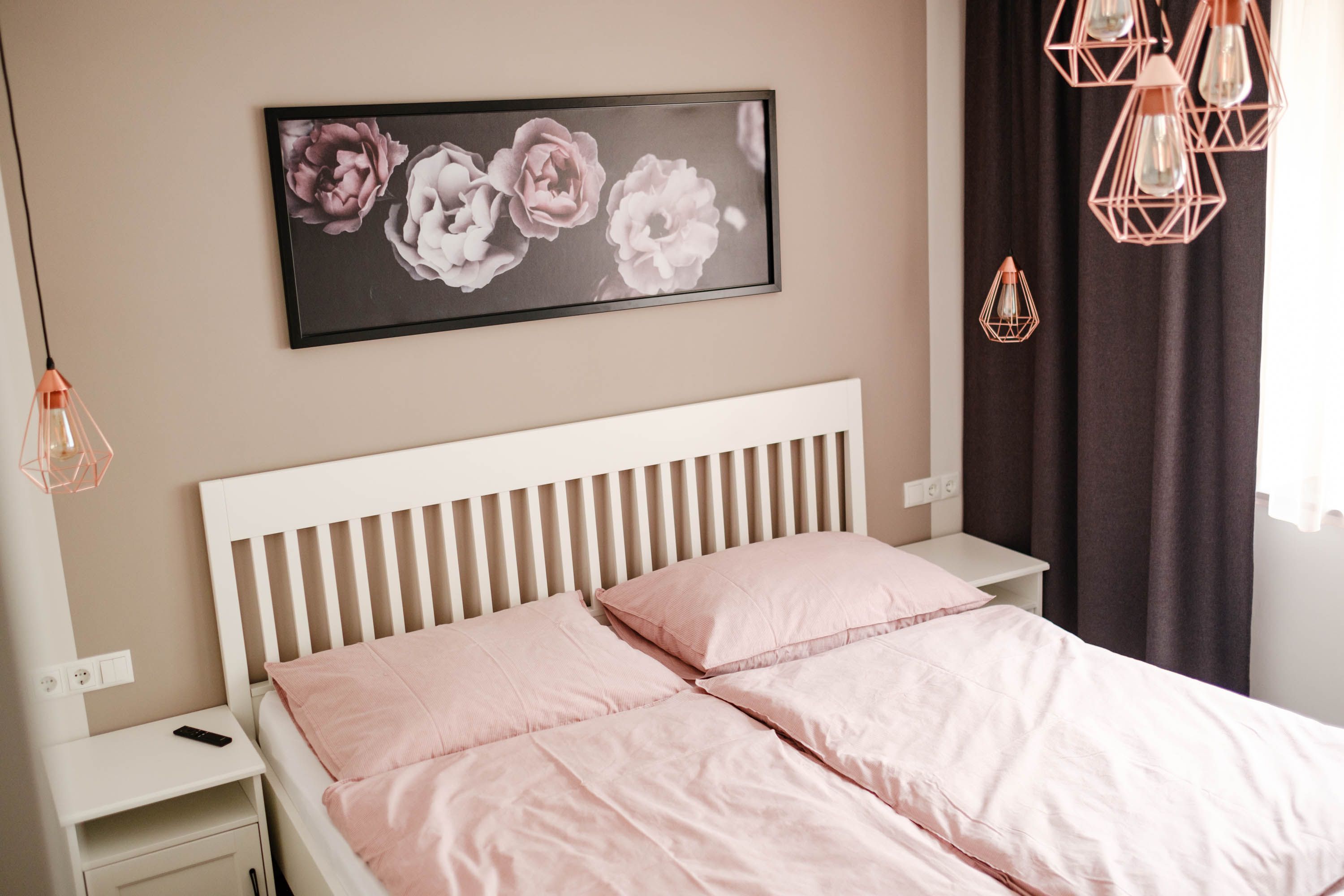 Schlafzimmer mit rosa Bettwäsche, weißem Bettgestell und Blumenbild an der Wand.