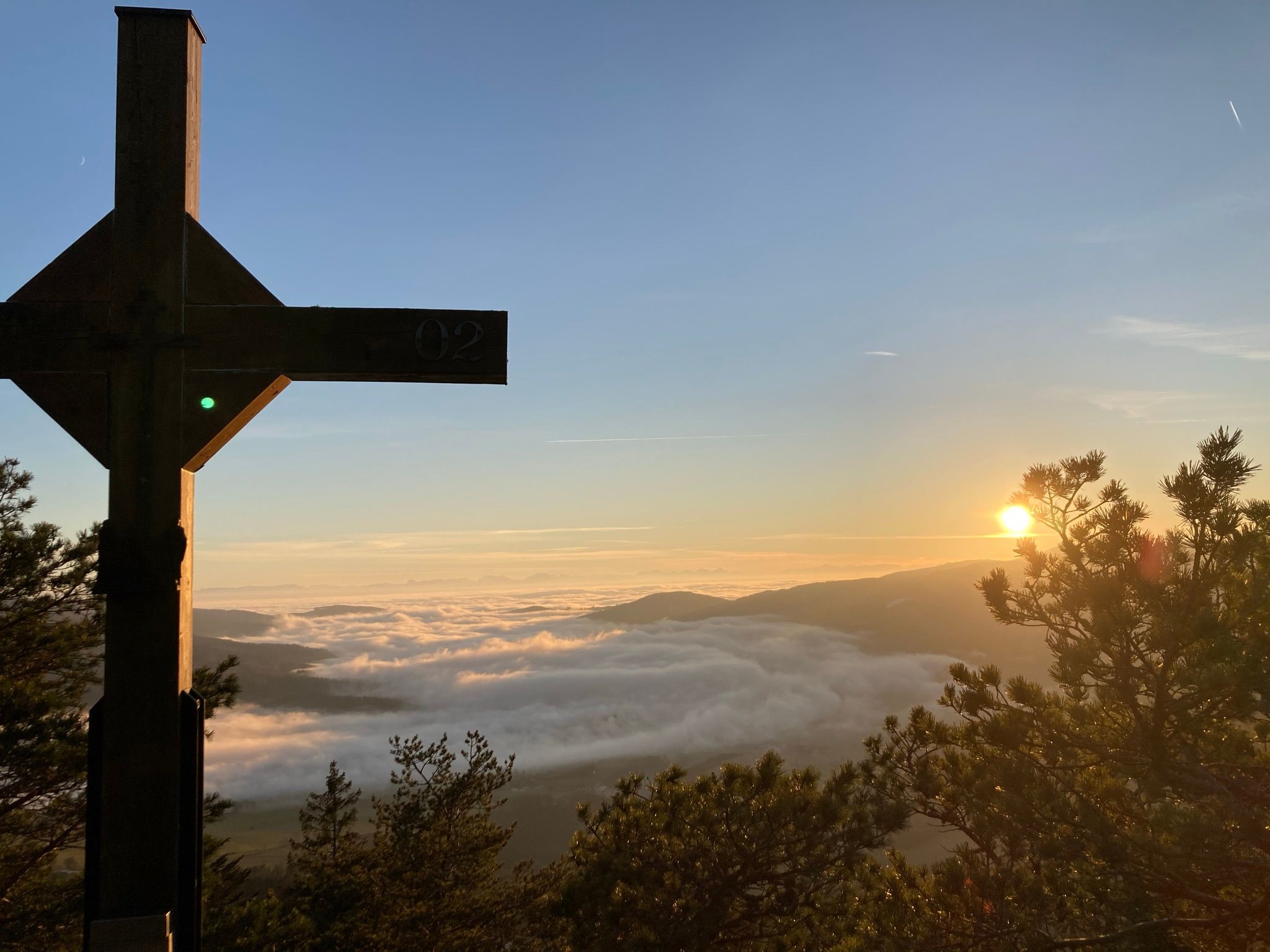 Ein Gipfelkreuz auf einem Berg mit Blick auf ein Nebelmeer und einen Sonnenaufgang.