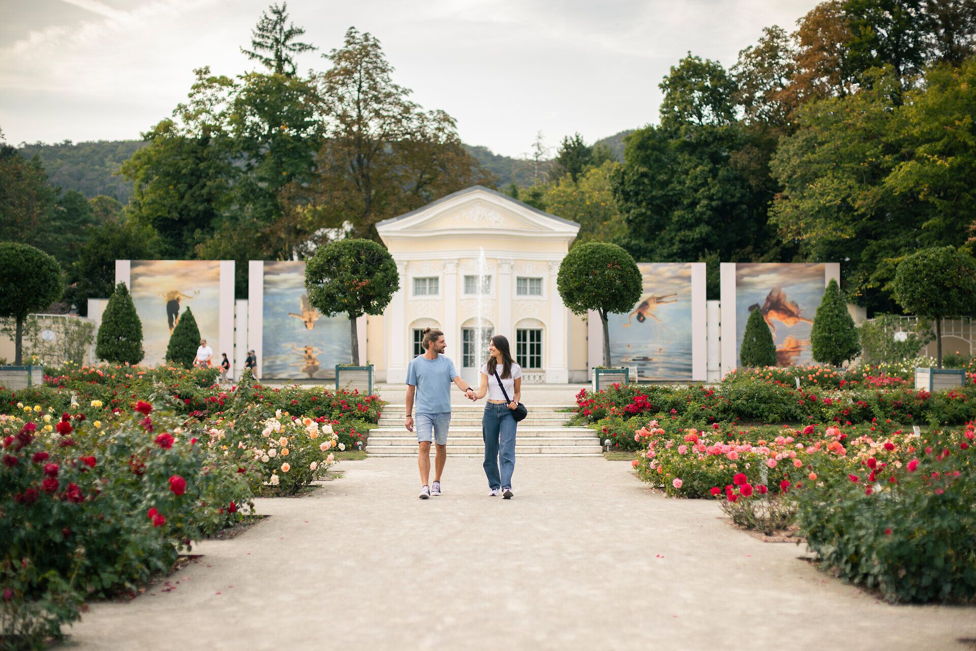 Zwischen blühenden Rosen im Doblhoffpark Baden spazieren eine Frau und ein Mann, vertieft in ein Gespräch. Im Hintergrund erhebt sich die eindrucksvolle Fotoausstellung von La Gacilly Baden Photo Baden, deren große Bilder das Szenario bereichern.