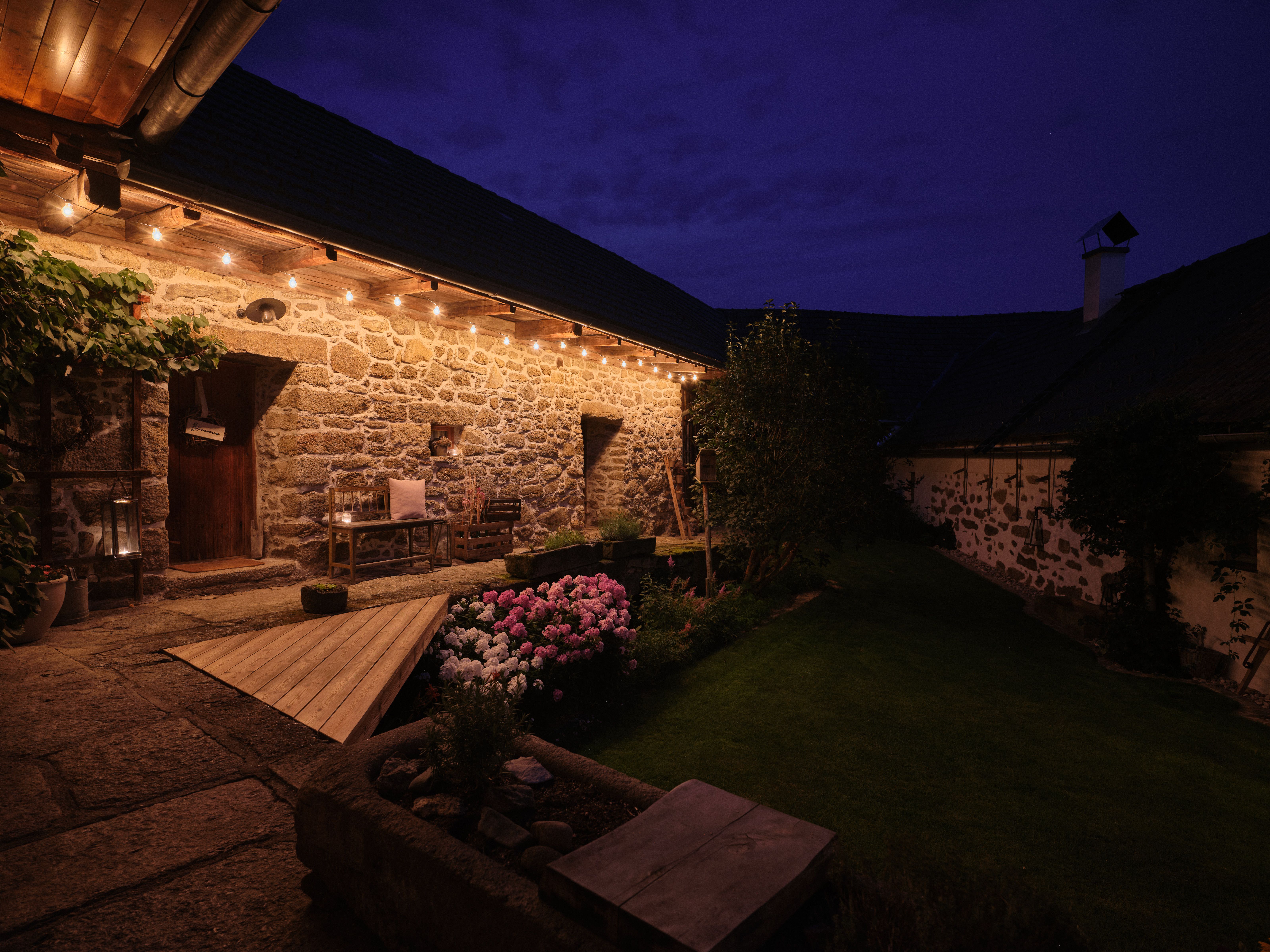 Ein beleuchteter Innenhof mit Steinmauern und Blumenbeeten bei Nacht.