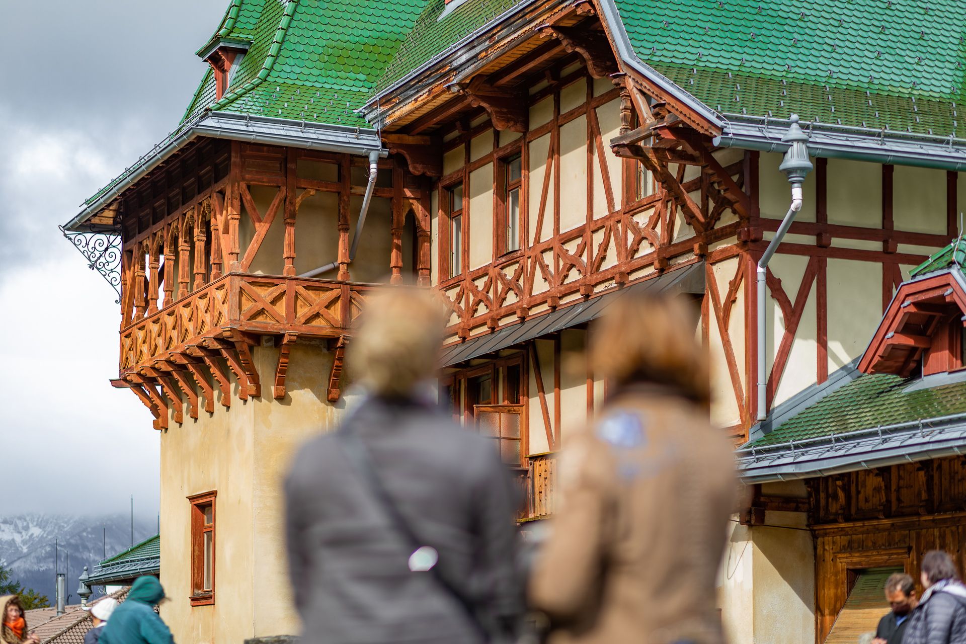 Historisches Gebäude mit grünem Dach und Fachwerk, unscharfe Personen im Vordergrund.