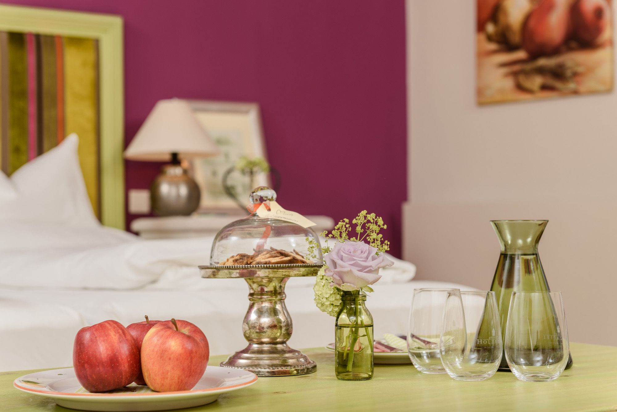 Ein stilvoll eingerichtetes Zimmer mit einem Tisch, auf dem Äpfel, Kekse unter einer Glasglocke, eine Vase mit Blume und Gläser stehen.