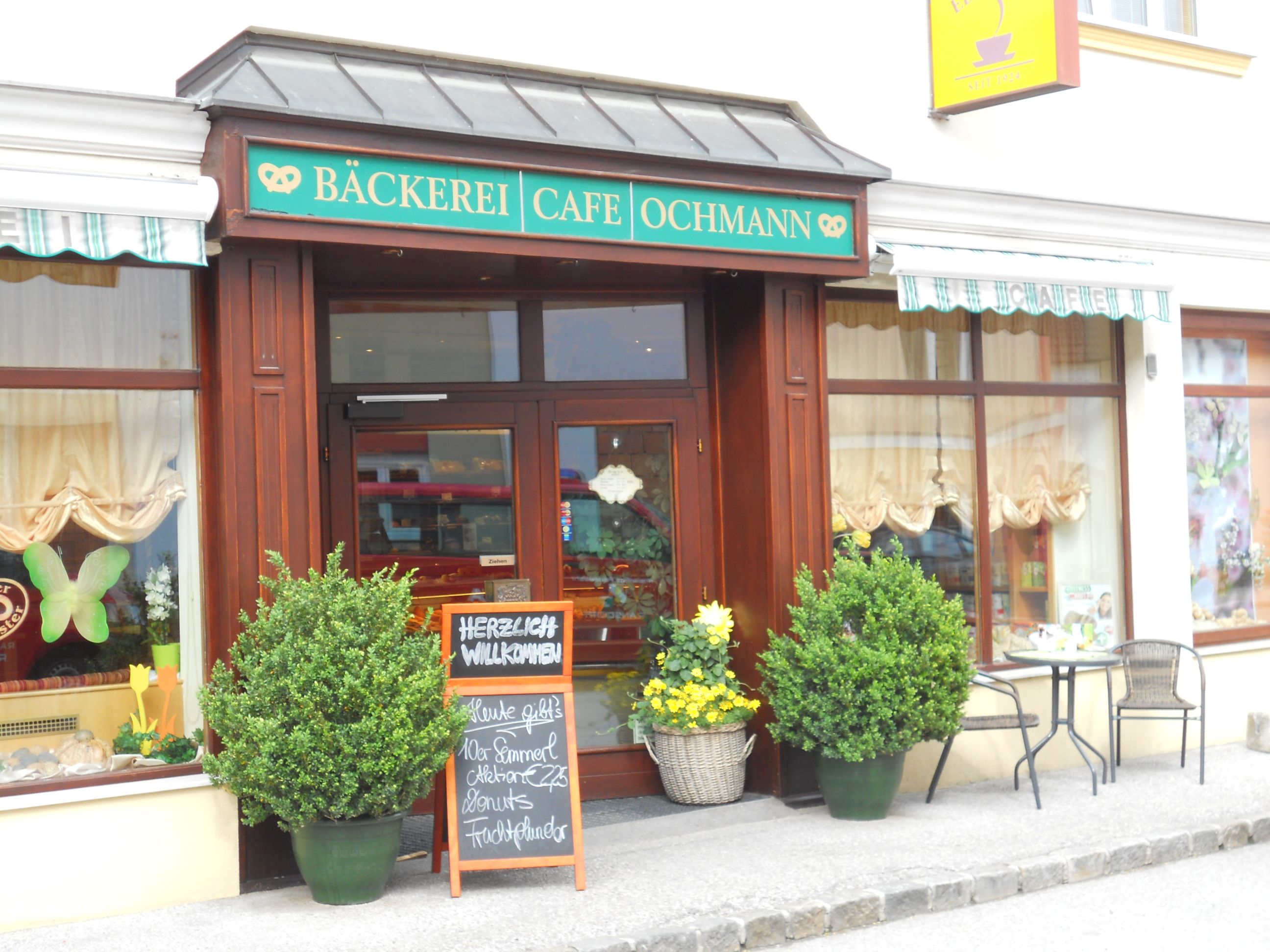 Eingang einer Bäckerei, dekoriert mit Pflanzen einem Schild und einem kleinen Tisch mit Stühlen.