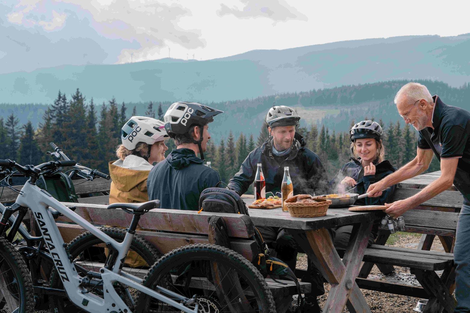 Gruppe von Mountainbikern sitzt an einem Holztisch in den Bergen und genießt eine Mahlzeit.