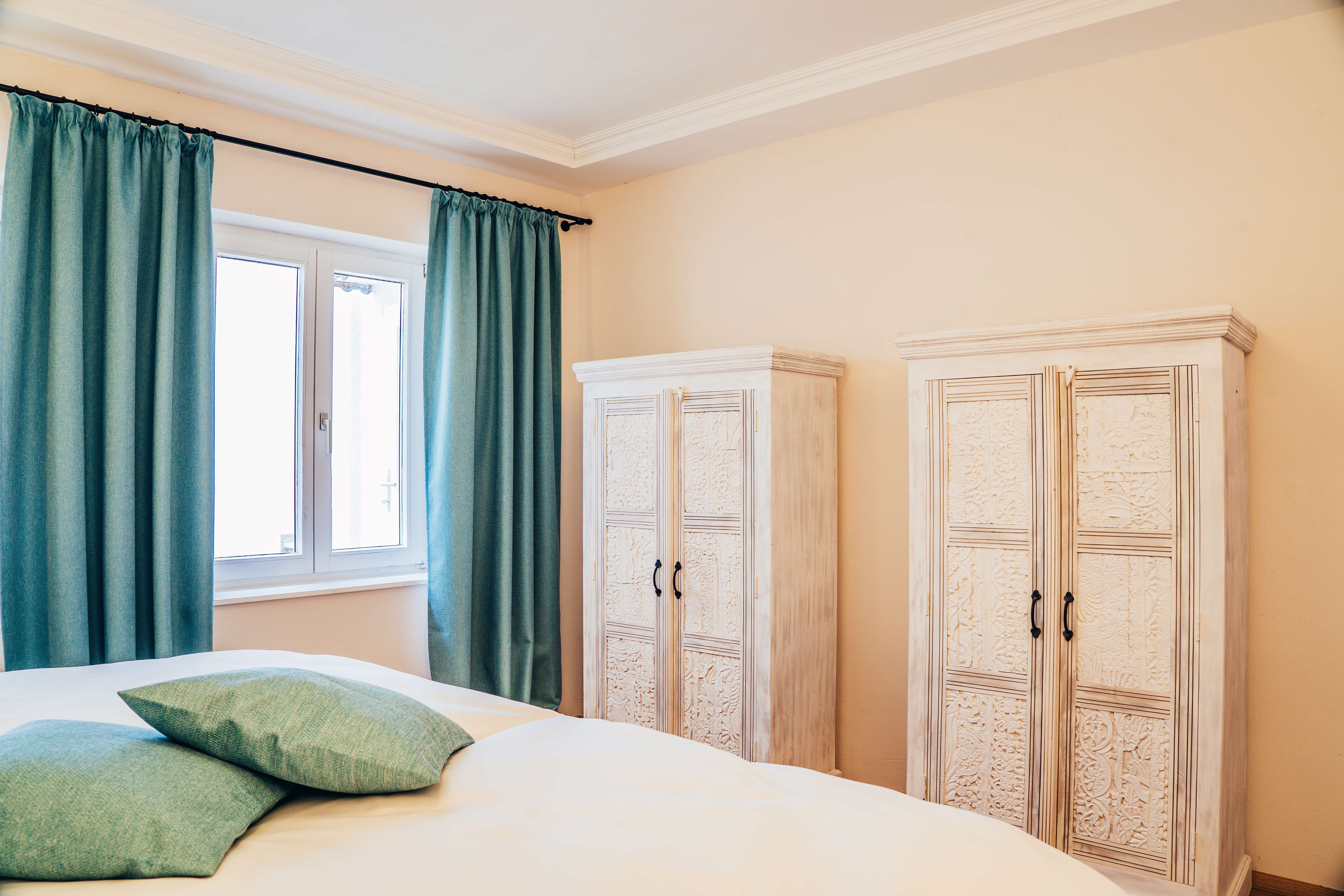 Schlafzimmer mit zwei weißen Schränken, türkisfarbenen Vorhängen und Kissen.