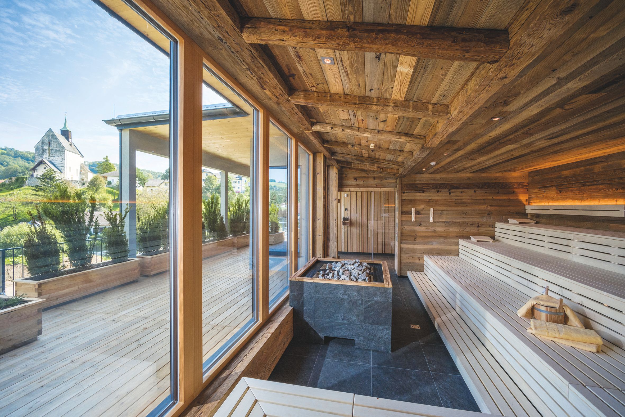 Innenansicht einer Sauna mit Holzbänken und großen Fenstern, die auf eine Terrasse und eine Kirche im Hintergrund blicken.