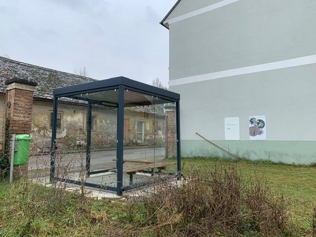 Eine moderne Bushaltestelle mit Glaswänden neben einem alten Gebäude und einer grünen Wiese.