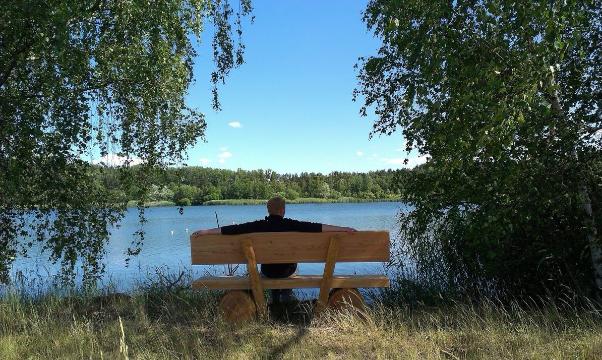Person sitzt auf einer Bank am Ufer eines Sees, umgeben von Bäumen.