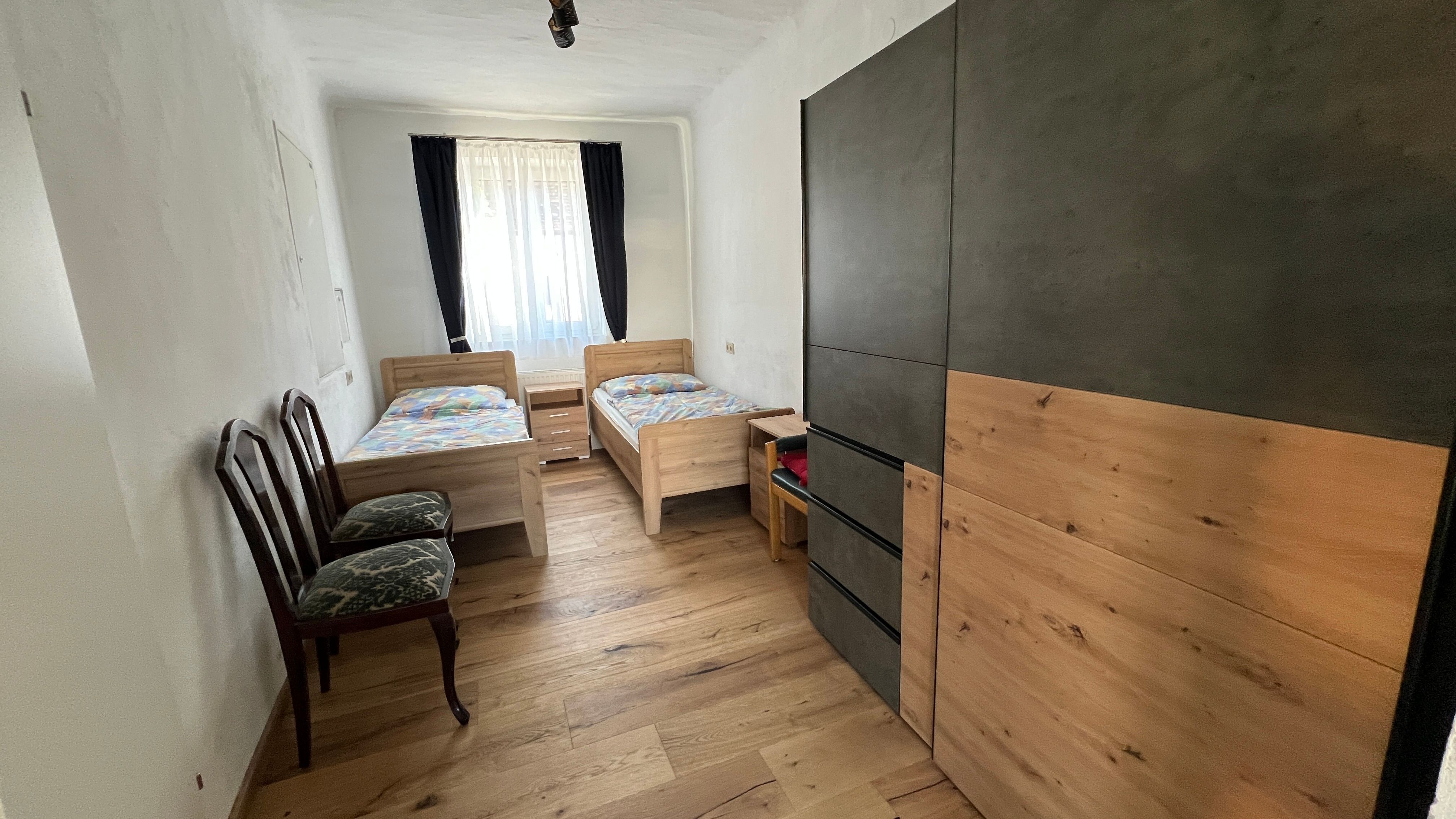 Ein kleines Schlafzimmer mit zwei Einzelbetten, einem Schrank und zwei Stühlen. Ein Fenster mit Vorhängen lässt Licht herein.