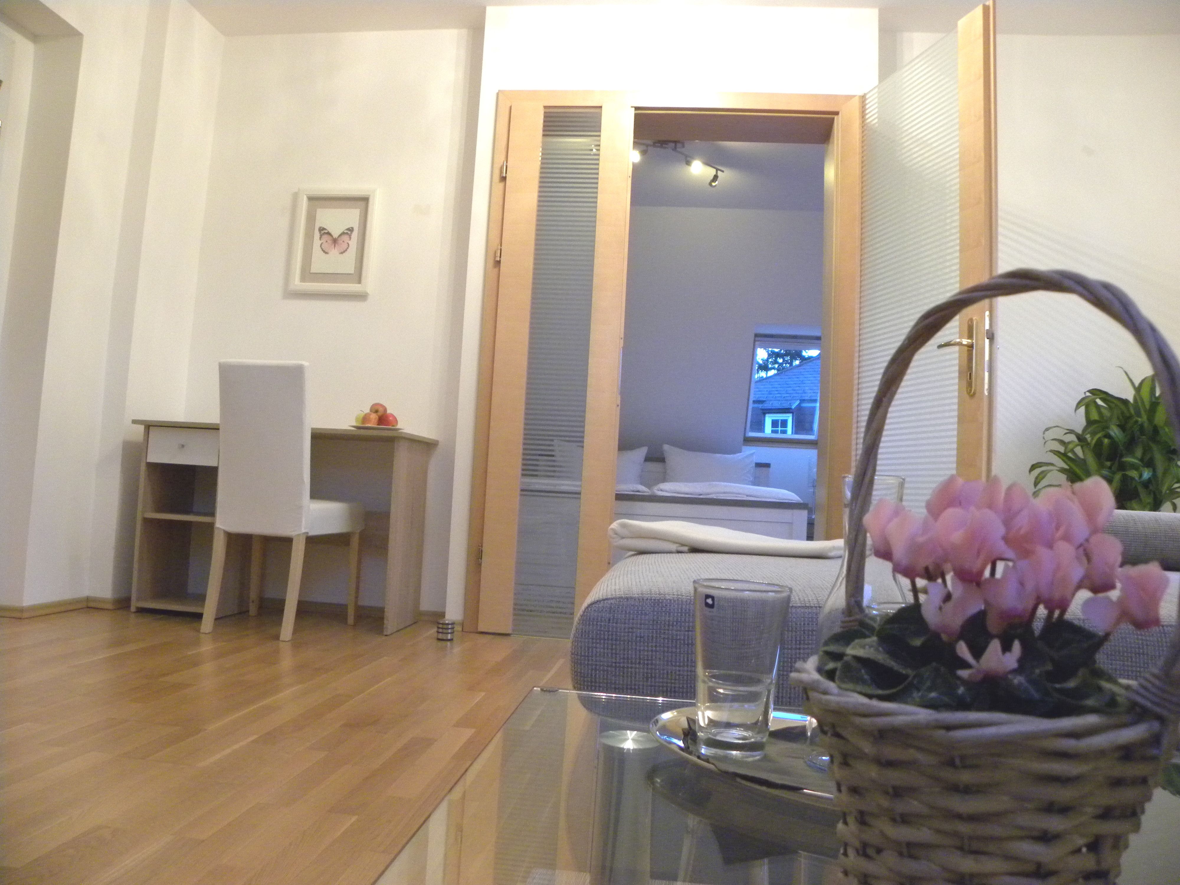 Innenraum mit Schreibtisch, Stuhl, Blumen im Korb und Blick ins Schlafzimmer.