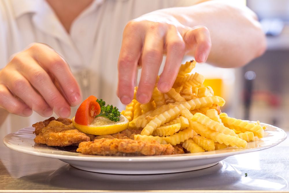 Ein Teller mit Wiener Schnitzel, Pommes frites, Zitrone und Tomate wird von einer Person angerichtet.