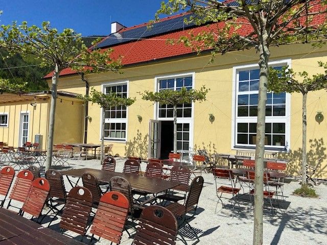 Außenbereich eines Restaurants mit gelbem Gebäude, roten Stühlen und Tischen unter Bäumen.