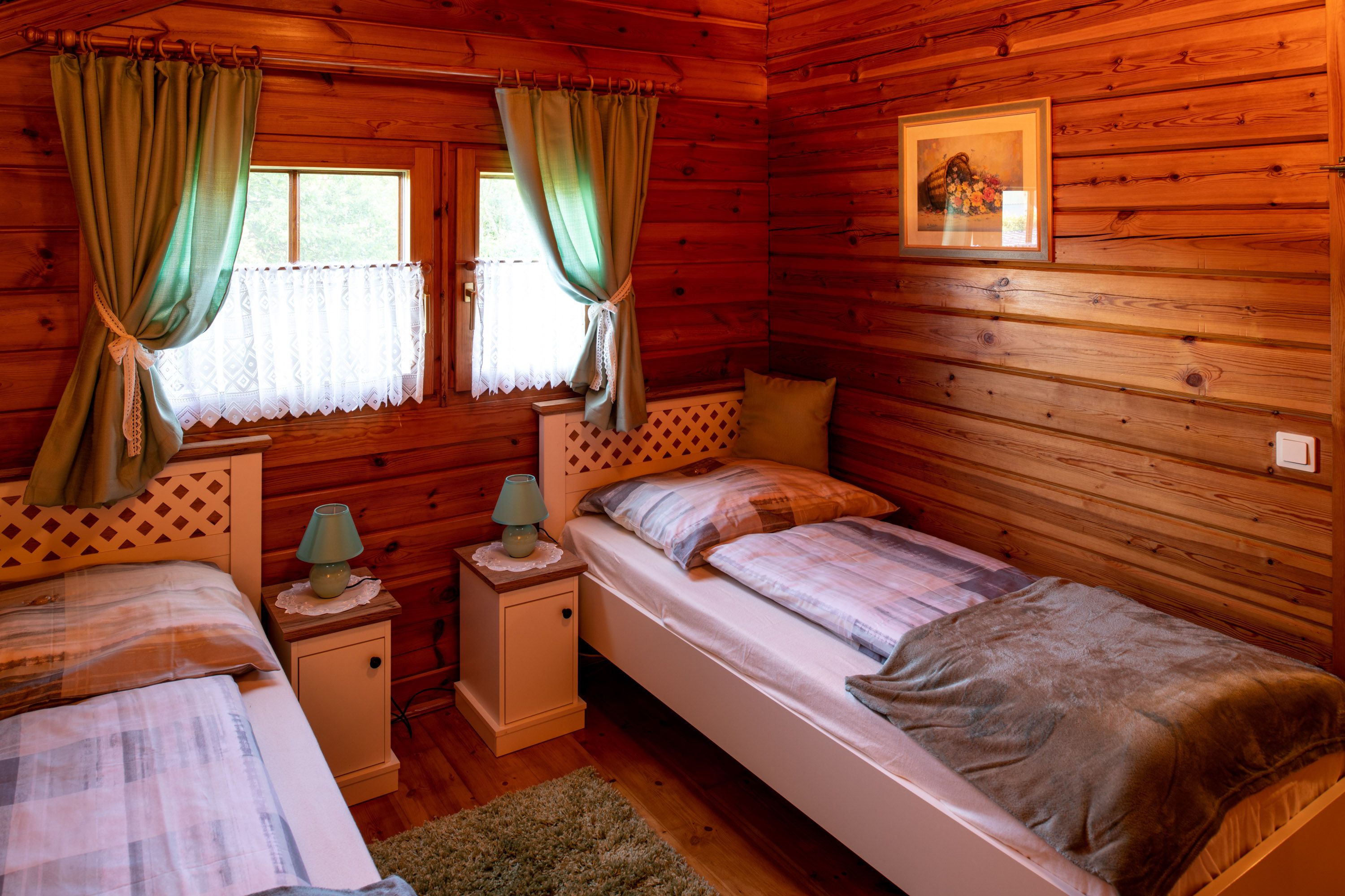 Gemütliches Schlafzimmer mit Holzwänden, zwei Einzelbetten, grünen Vorhängen und Nachttischen mit Lampen.