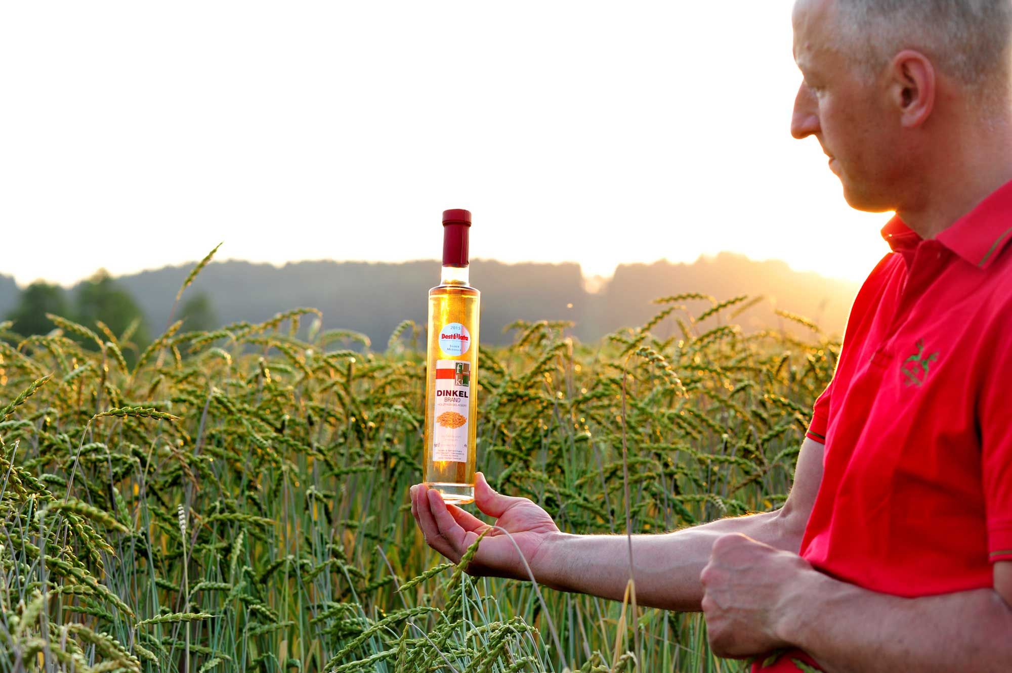 Ein Mann in rotem Hemd hält eine Flasche Dinkelbrand in einem Getreidefeld bei Sonnenuntergang.