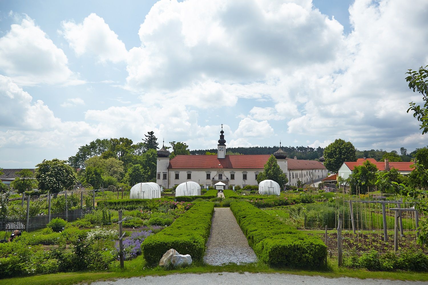 Ein gepflegter Garten mit Gewächshäusern vor einem historischen Gebäude mit rotem Dach und Turm, umgeben von Bäumen und Wolken am Himmel.