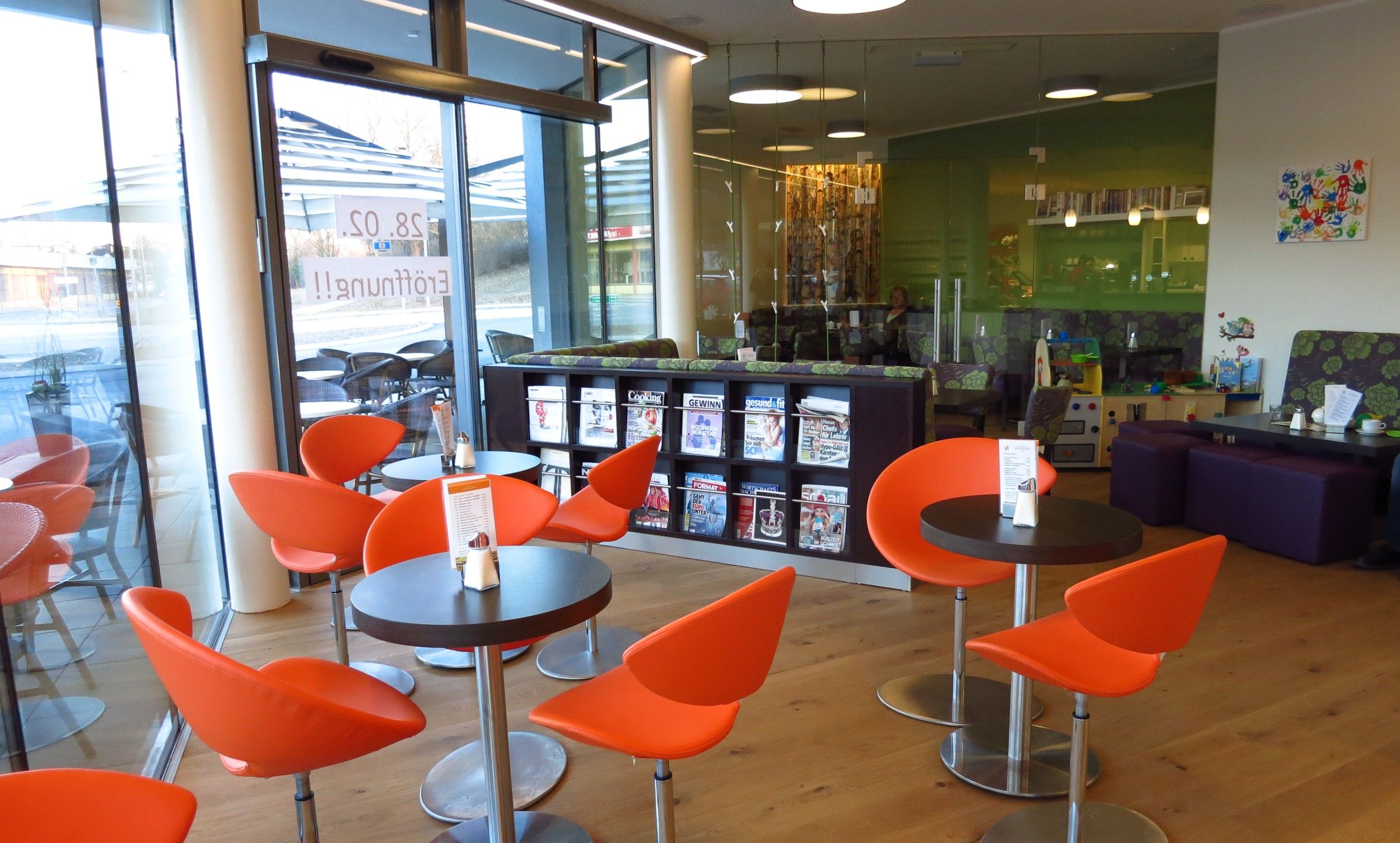 Innenansicht eines modernen Cafés mit orangefarbenen Stühlen und Zeitschriftenregal.