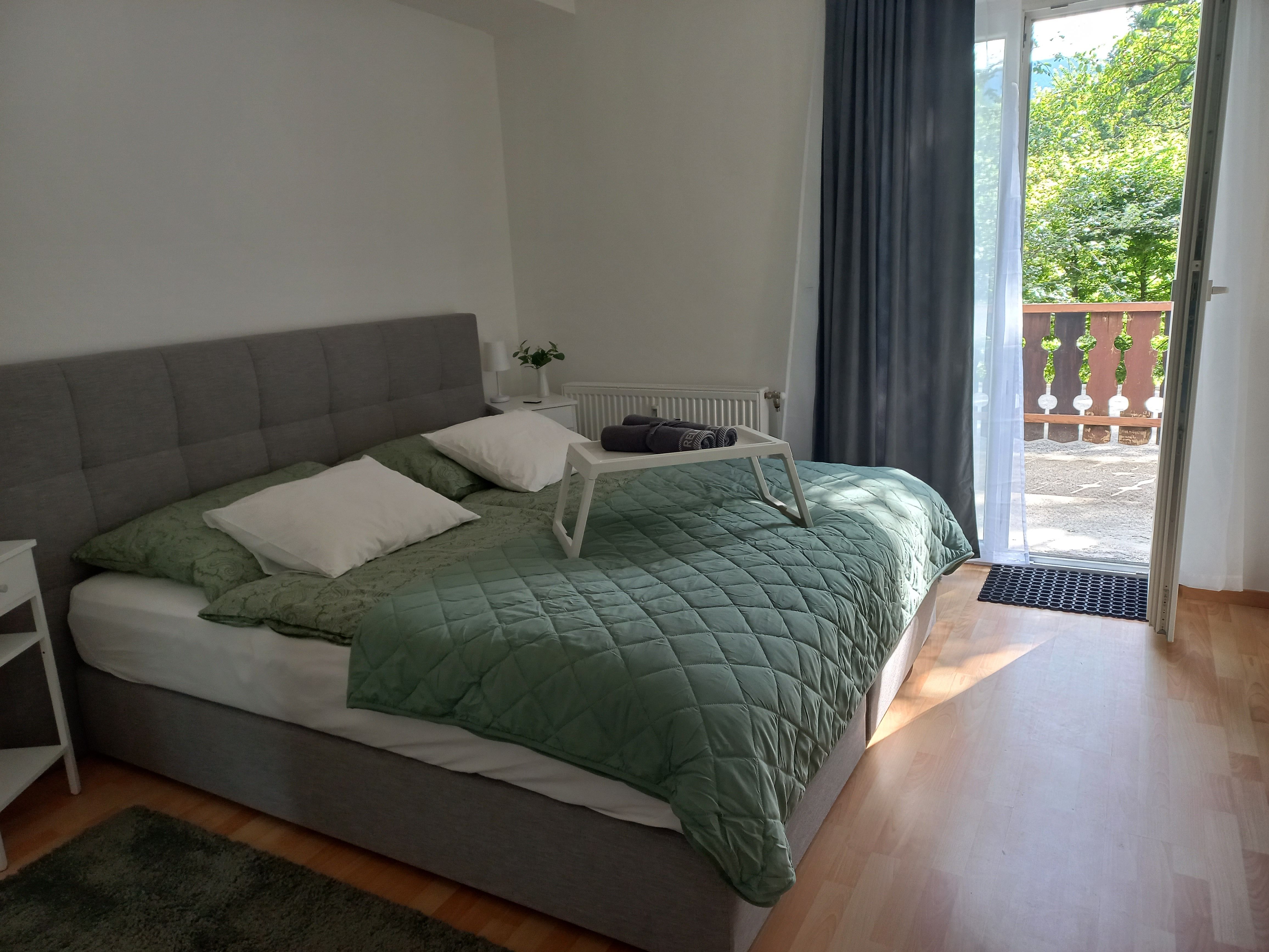 Ein gemütliches Schlafzimmer mit einem grauen Bett, grüner Bettdecke und offenem Balkon mit Blick ins Grüne.