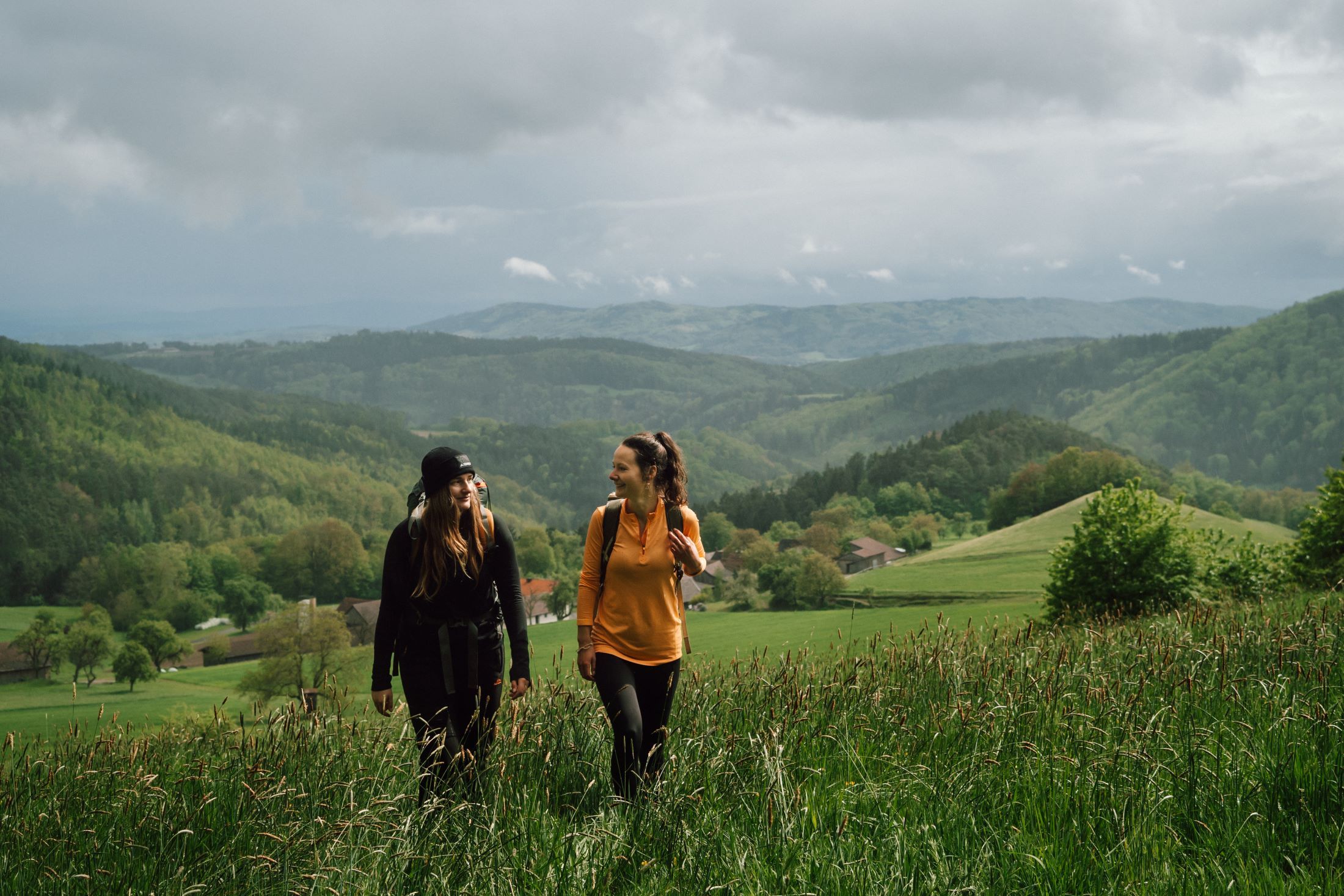 Zwei Frauen wandern durch eine grüne Landschaft mit Hügeln im Hintergrund.