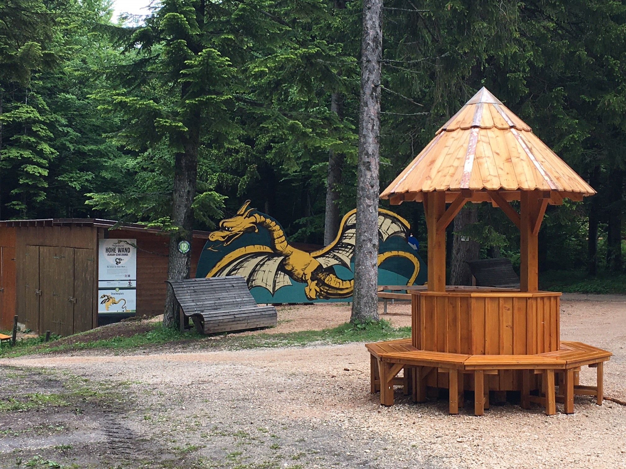 Ein hölzerner Pavillon auf einem Waldplatz mit einem gemalten Drachen im Hintergrund.