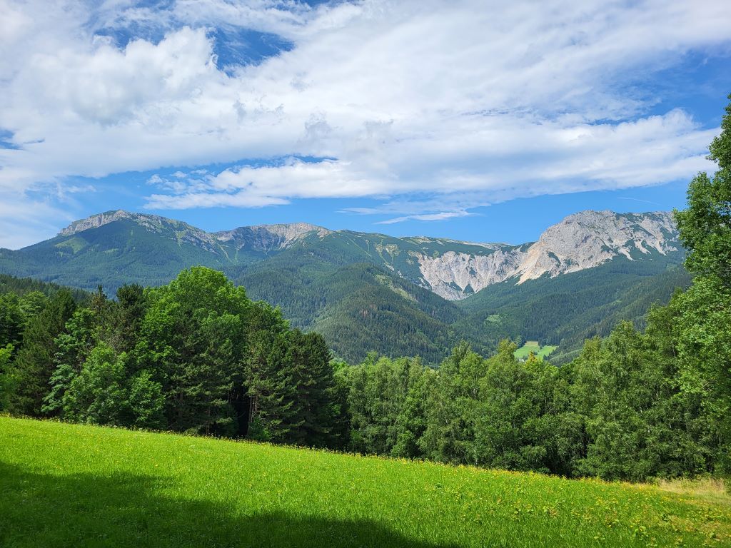 Berglandschaft mit grünen Wiesen und bewaldeten Hügeln unter blauem Himmel.