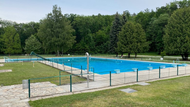 Ein leeres Freibad mit einem großen Schwimmbecken, umgeben von Bäumen und Rasenflächen.
