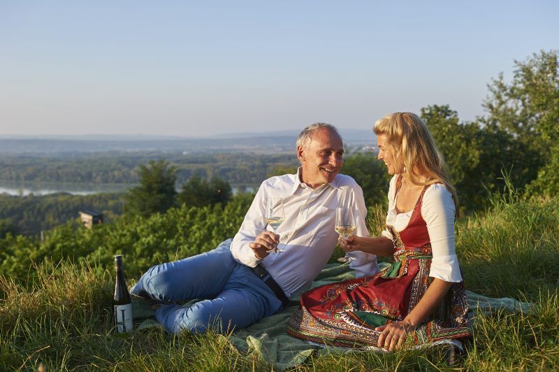 Ein Paar sitzt auf einer Wiese und trinkt Wein mit Blick auf eine Landschaft.