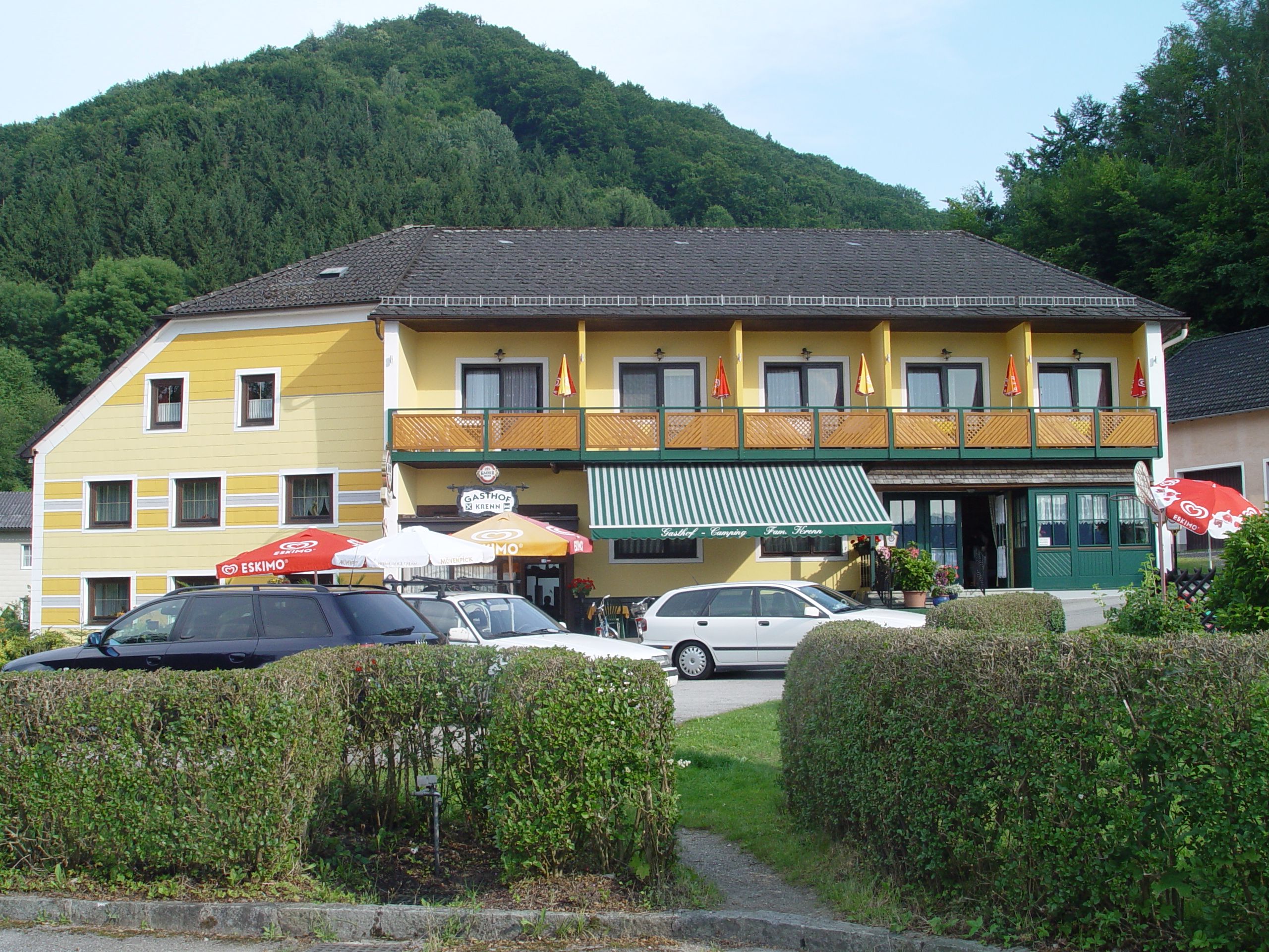 Gelbes Gasthaus mit Balkon und Sonnenschirmen vor einem bewaldeten Hügel.