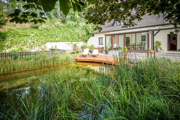 Ein Haus mit Terrasse und Schwimmbiotop im Garten, umgeben von üppiger Vegetation.
