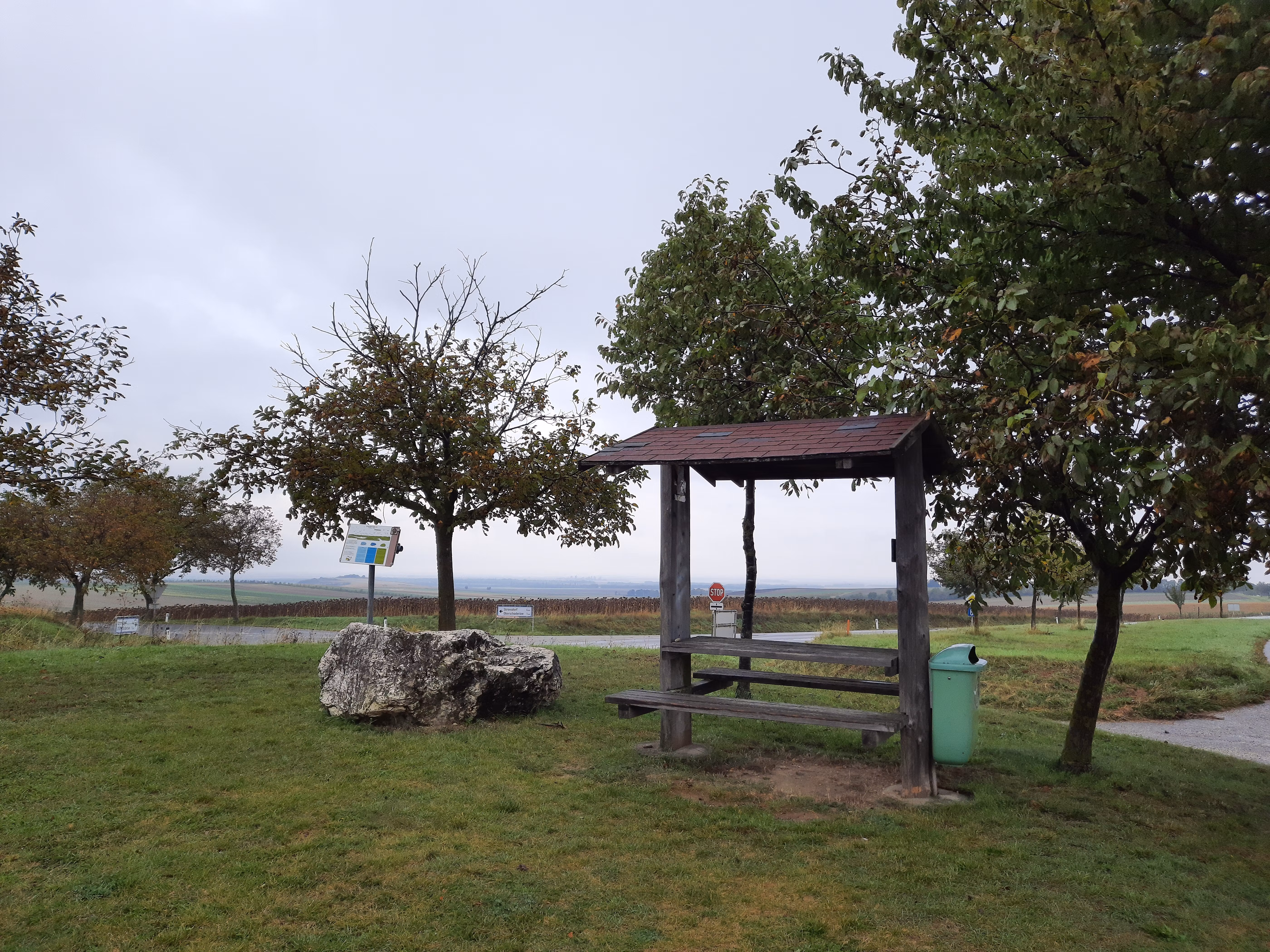 Ein Rastplatz mit Holzbänken und einem Tisch, umgeben von Bäumen und einer Wiese, unter einem bewölkten Himmel.