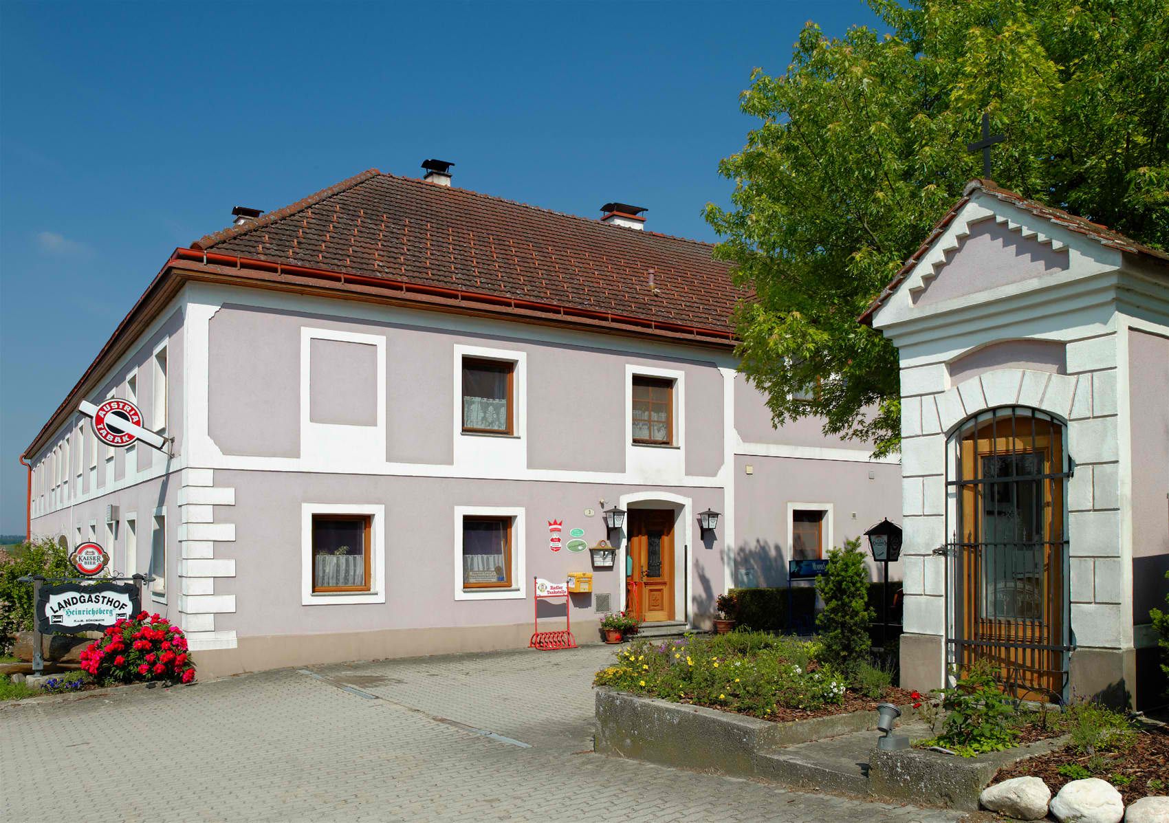 Ein traditioneller Landgasthof mit grauer Fassade und roten Dachziegeln, umgeben von Blumen und Bäumen.