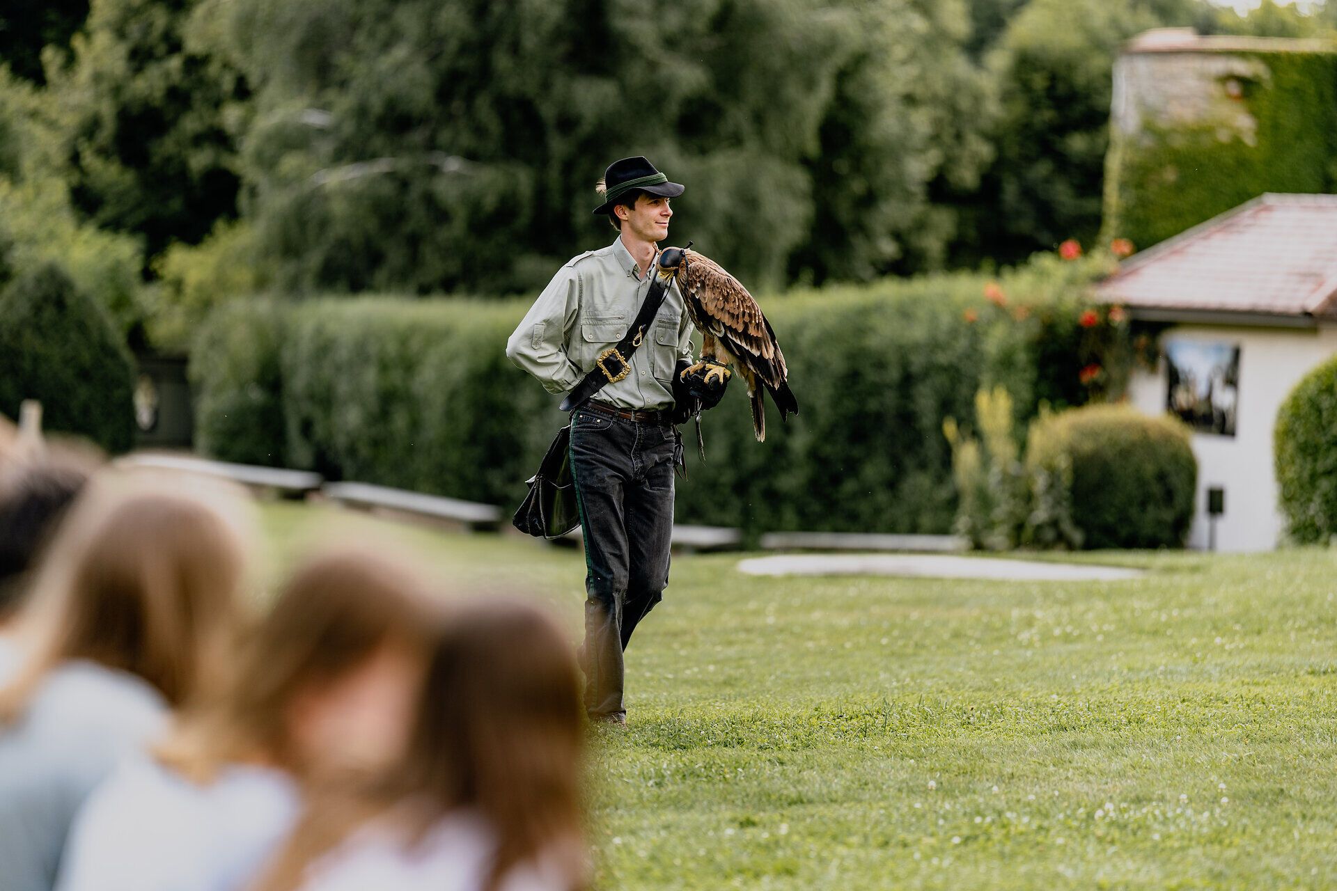 Ein Falkner mit einem Greifvogel auf dem Arm steht auf einer Wiese vor einer Gruppe von Zuschauern.