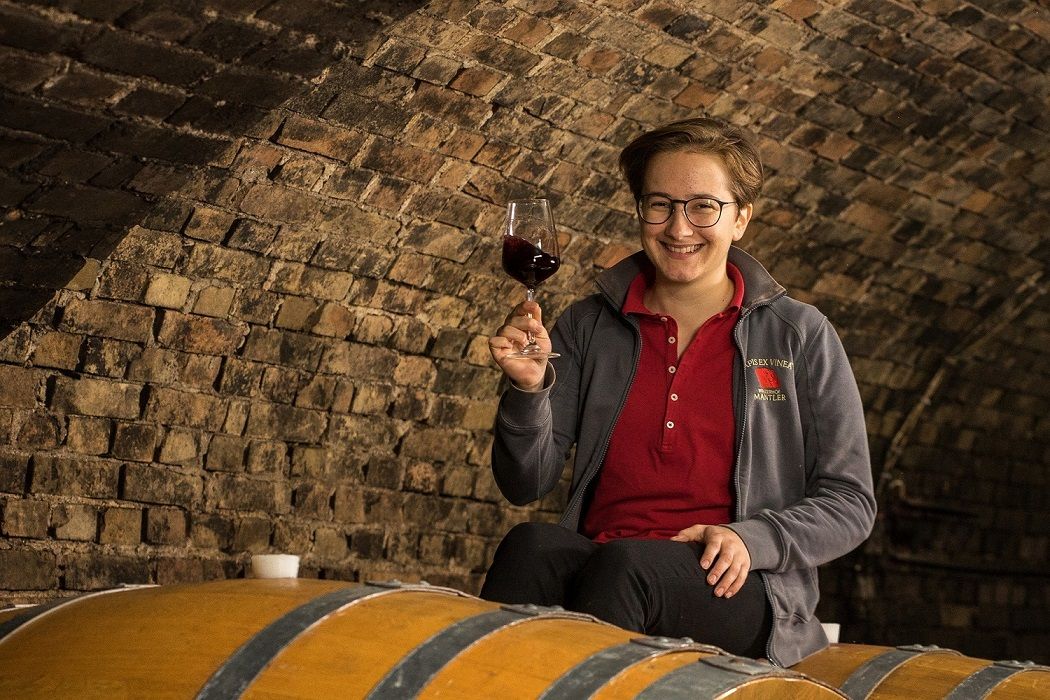 Eine Person sitzt auf einem Weinfass in einem Weinkeller und hält ein Glas Rotwein in der Hand.