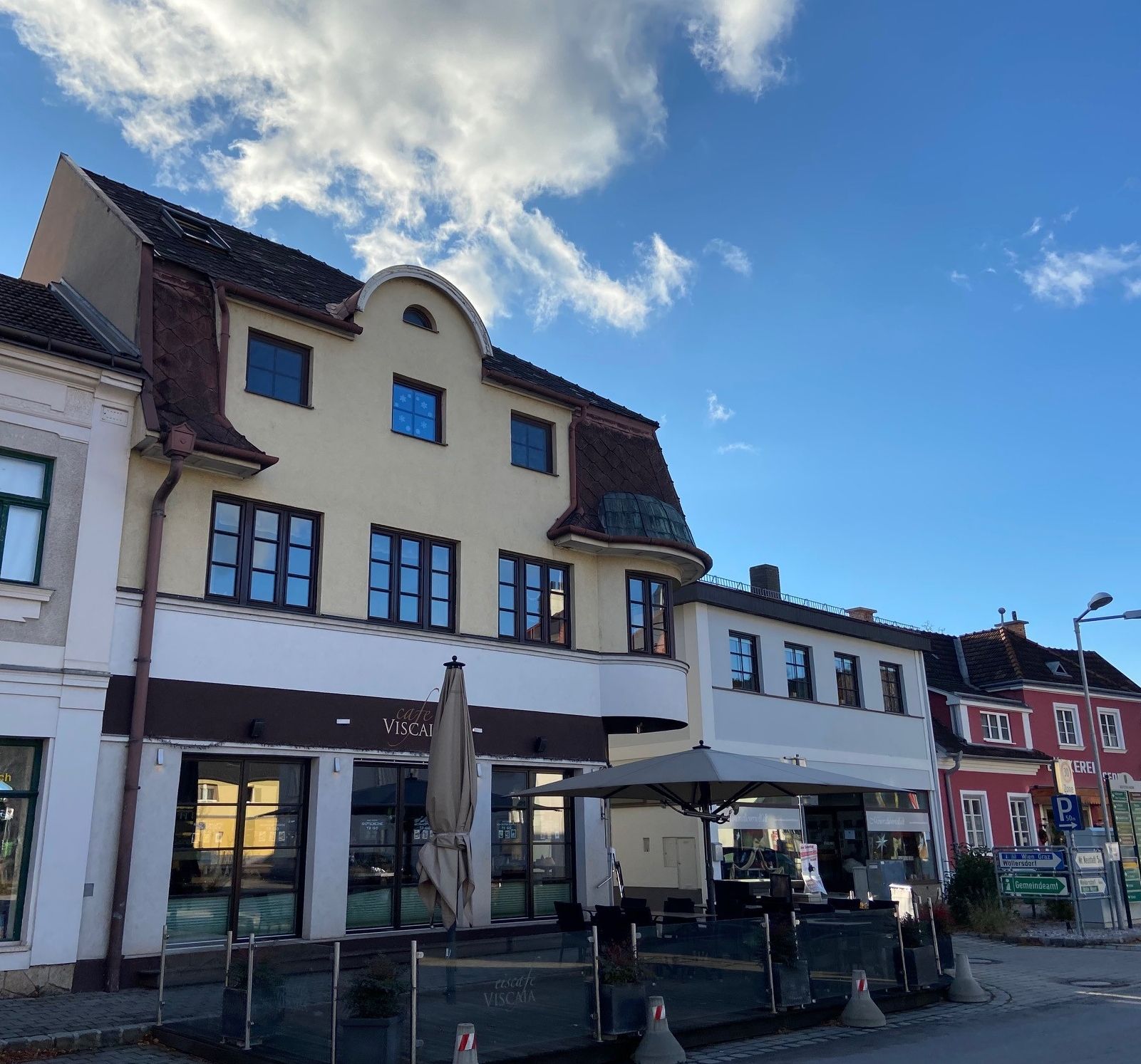 Außenansicht eines Cafés mit Terrasse und Sonnenschirm vor einem mehrstöckigen Gebäude.