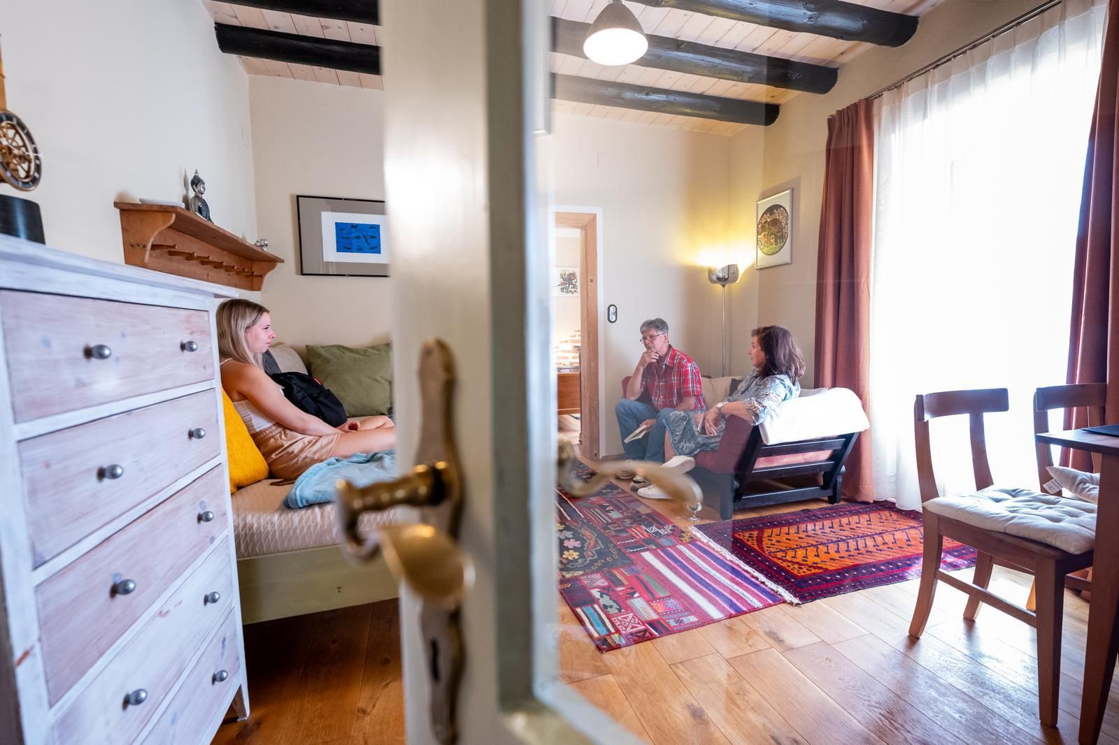 Drei Personen sitzen in einem gemütlichen Wohnzimmer mit Holzboden und buntem Teppich.