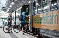 Ein Radfahrer steigt mit seinem Mountainbike in die wartende Bahn ein, während ein weiterer Radler auf dem Bahnsteig steht. Die Atmosphäre ist lebhaft und einladend, perfekt für Abenteuerlustige, die die malerische Landschaft des Waldviertels erkunden möchten.