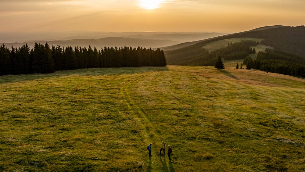 Drei Personen wandern über eine grüne Wiese bei Sonnenaufgang, umgeben von Wäldern und Hügeln.
