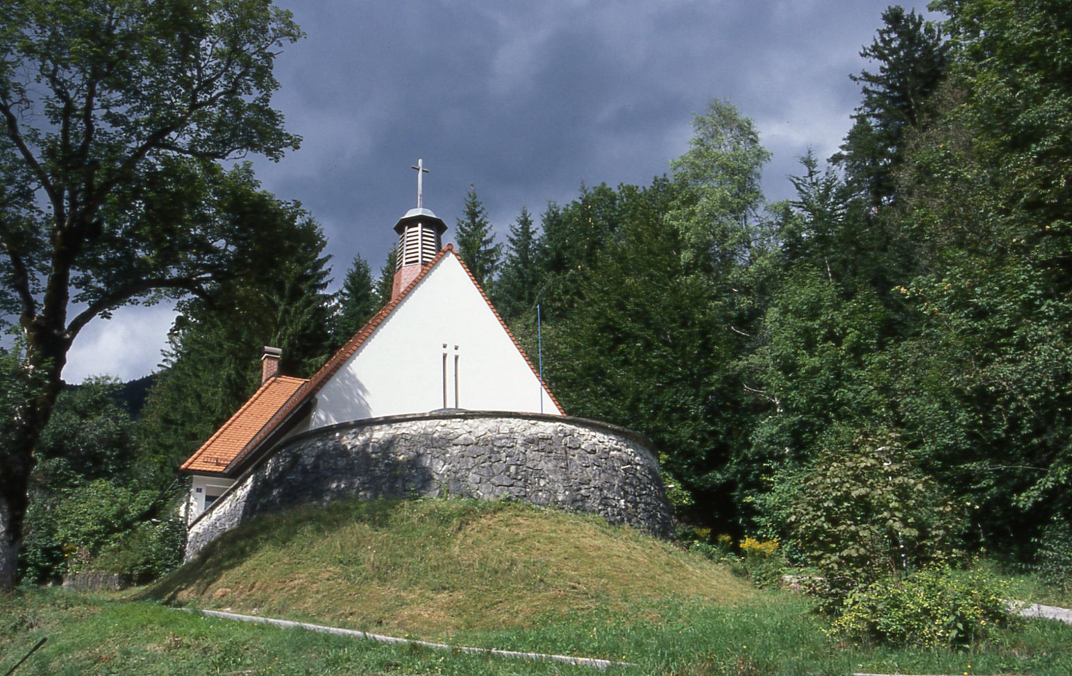 Kleine Kirche im Wald mit steinernem Sockel und rotem Dach.