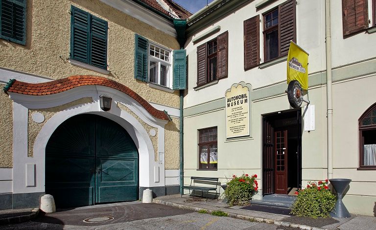 Eingang eines Automobilmuseums mit gelber Fahne und Pflanzen.