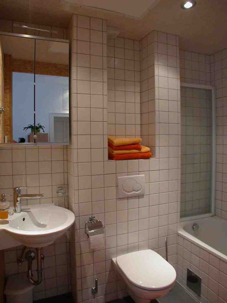 Modernes Badezimmer mit weißen Fliesen, Waschbecken, Toilette und Badewanne. Handtücher in einem Regal.