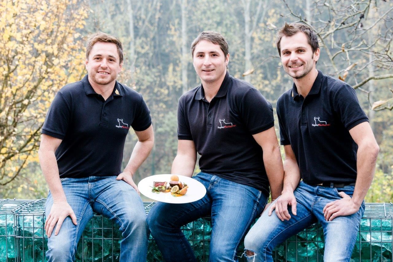 Drei Männer in schwarzen Poloshirts sitzen draußen und halten einen Teller mit Essen.
