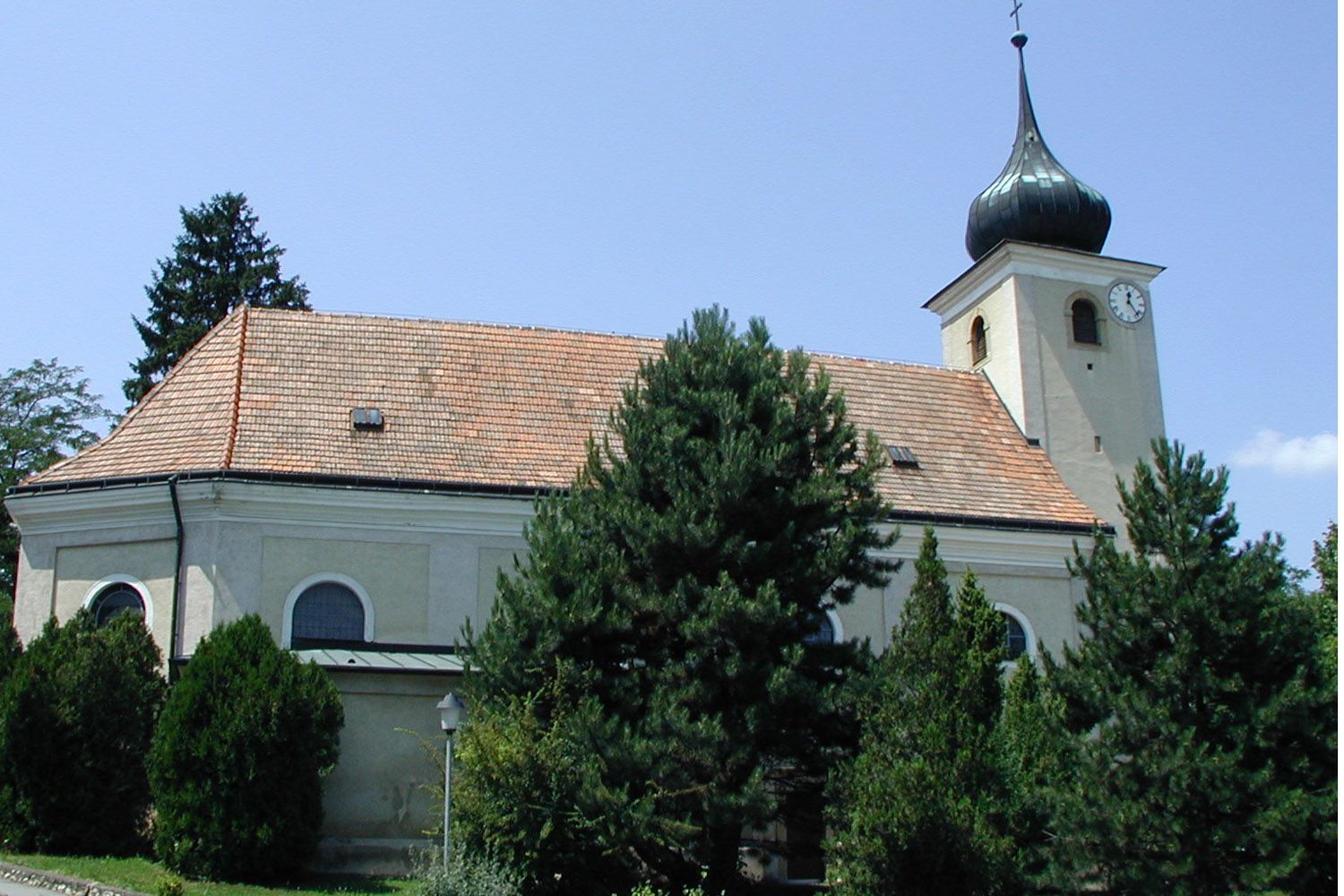 Kirche in Schleinbach mit Turm und Uhr, umgeben von Bäumen.