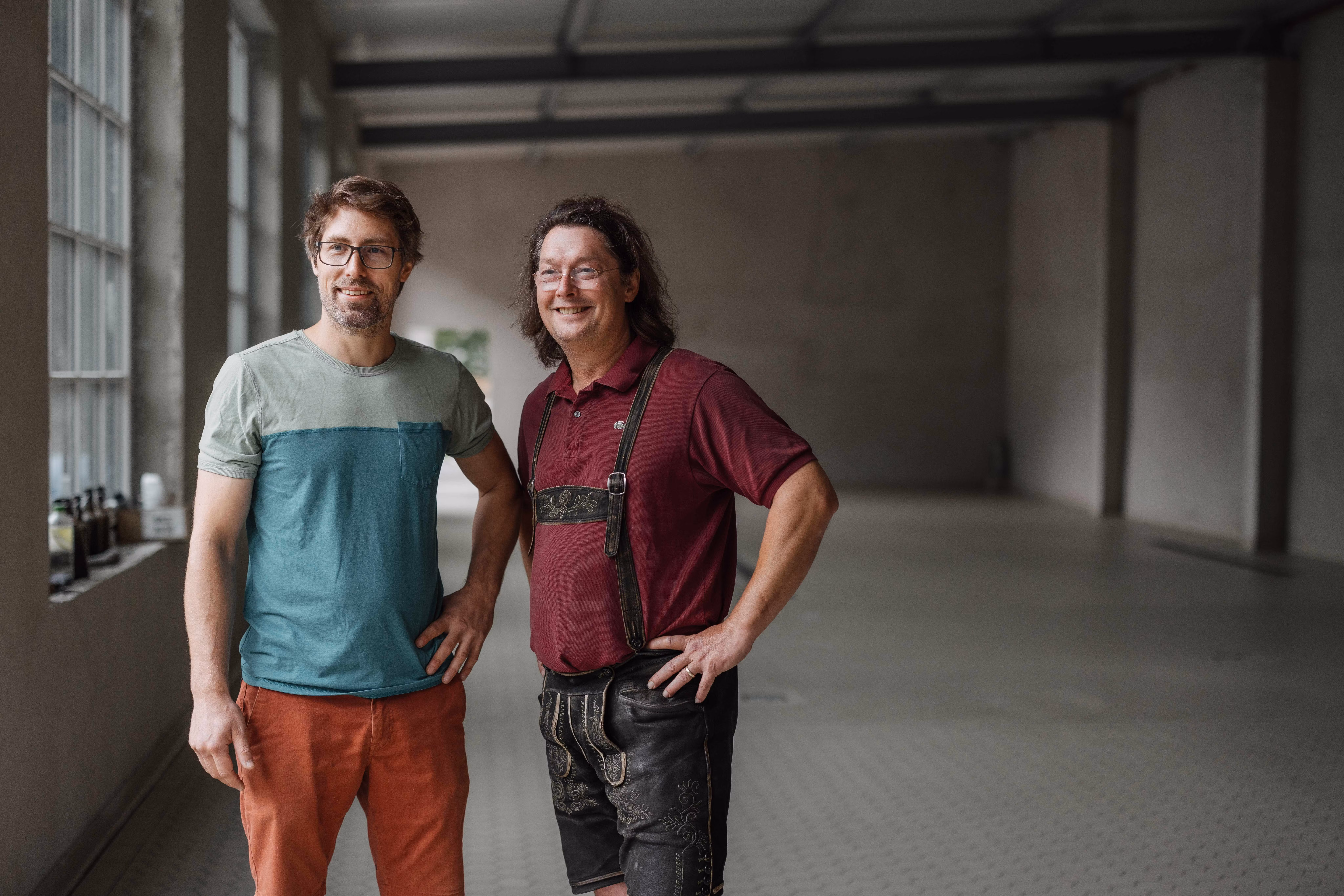 Zwei Männer, die Gründer von Raxbräu, stehen in einem leeren Raum, einer trägt ein T-Shirt und der andere Lederhosen.