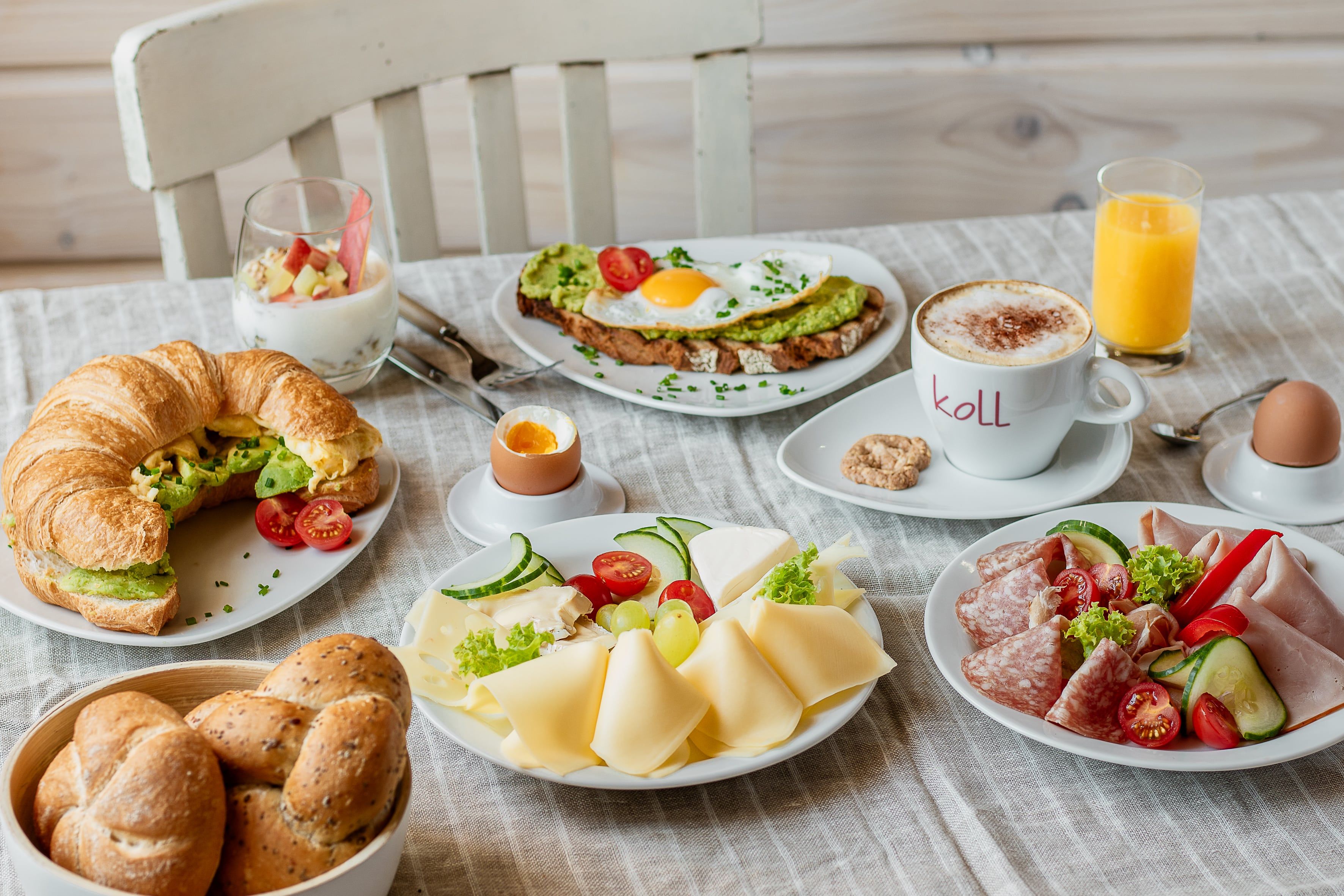 Ein reichhaltiges Frühstück mit Croissant, Brötchen, Käse, Aufschnitt, Avocado-Toast, Joghurt, Cappuccino, Orangensaft und weichgekochtem Ei.
