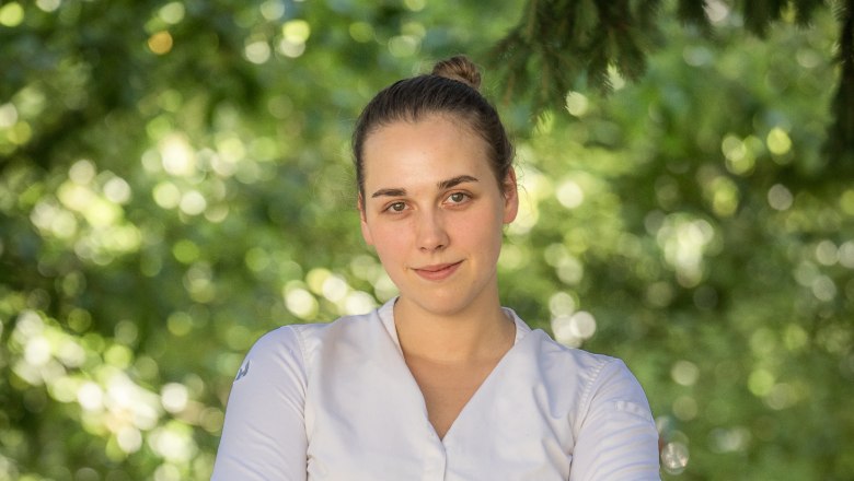 Eine Frau in weißem Hemd steht mit verschränkten Armen vor einem unscharfen, grünen Hintergrund.