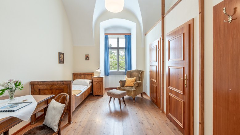 Einzelzimmer im Stift Altenburg mit Bett, Sessel, Tisch und Fenster mit blauen Vorh&auml;ngen.