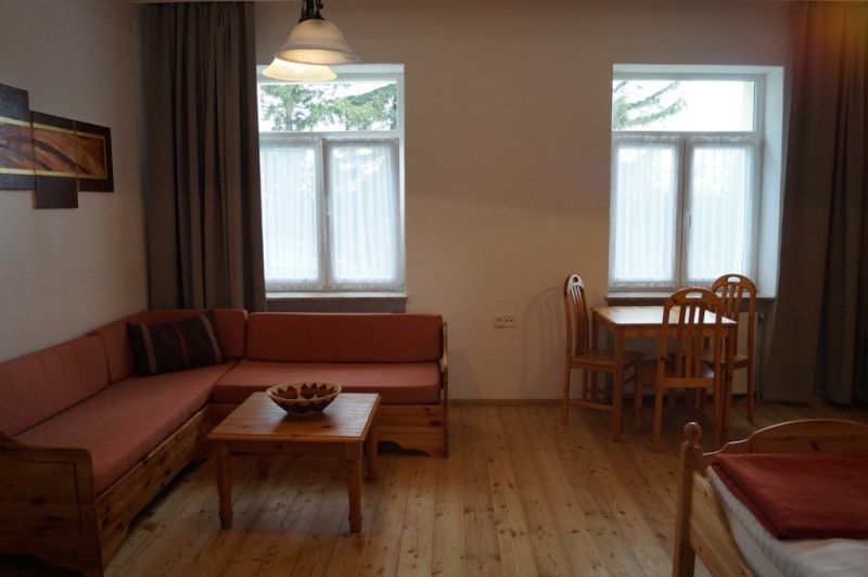Gemütliches Zimmer mit Holzmöbeln, Sofa, Tisch und Stühlen, zwei Fenstern und einem Bild an der Wand.
